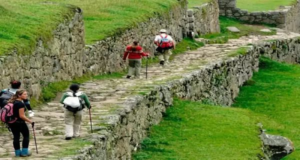 Turistas caminan por un sendero de piedra rodeado de muros incas y paisajes verdes, una experiencia cl�sica del turismo de aventura en Cusco.